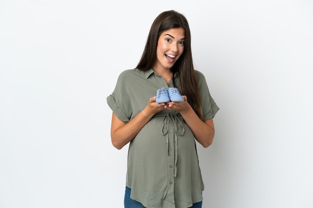 Foto jovem brasileira isolada em um fundo branco grávida e segurando botinhas de bebê com expressão de surpresa