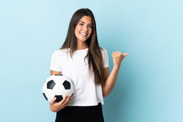 Jovem brasileira jogadora de futebol isolada em parede azul apontando para o lado para apresentar um produto