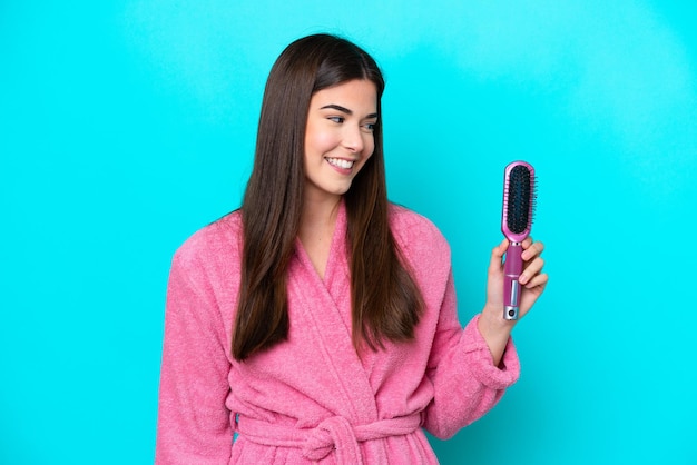 Jovem brasileira segurando escova de cabelo isolada em fundo azul com expressão feliz