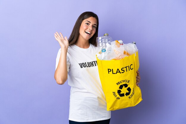 Jovem brasileira segurando uma sacola cheia de garrafas de plástico para reciclar isolada na parede roxa saudando com a mão com expressão feliz