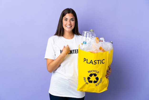 Jovem brasileira segurando uma sacola cheia de garrafas plásticas para reciclar isolada na parede roxa com expressão facial surpresa