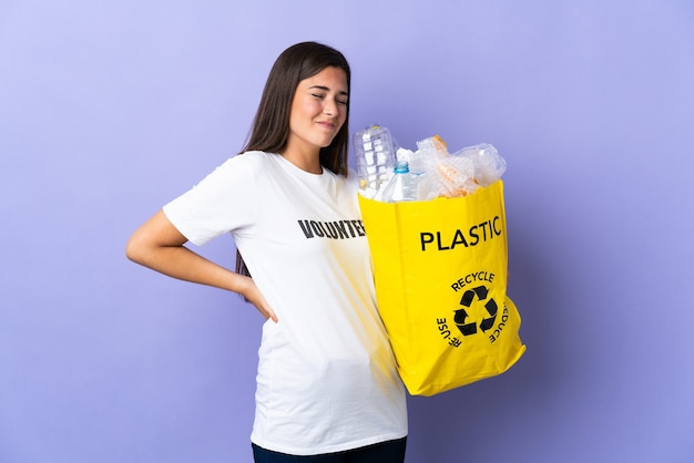 Jovem brasileira segurando uma sacola cheia de garrafas plásticas para reciclar isolada na parede roxa sofrendo de dor nas costas por ter feito um esforço