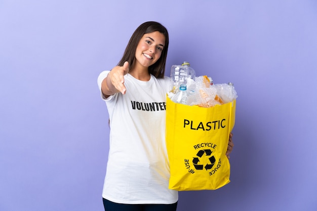 Jovem brasileira segurando uma sacola cheia de garrafas plásticas para reciclar isolada no roxo apertando as mãos para fechar um bom negócio