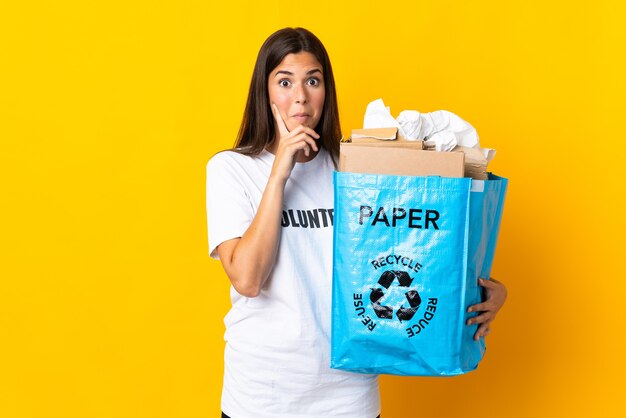 Jovem brasileira segurando uma sacola cheia de papel para reciclar isolada em um fundo amarelo pensando uma ideia