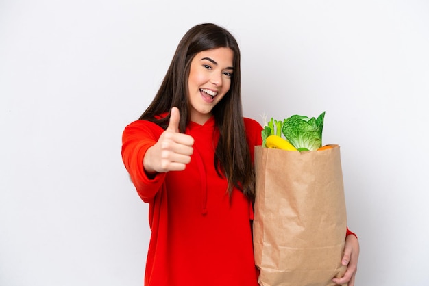 Jovem brasileira segurando uma sacola de compras isolada no fundo branco com polegares para cima porque algo bom aconteceu