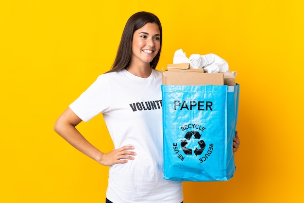 Jovem brasileira segurando uma sacola de reciclagem cheia de papel para reciclar em amarelo posando com os braços na cintura e sorrindo