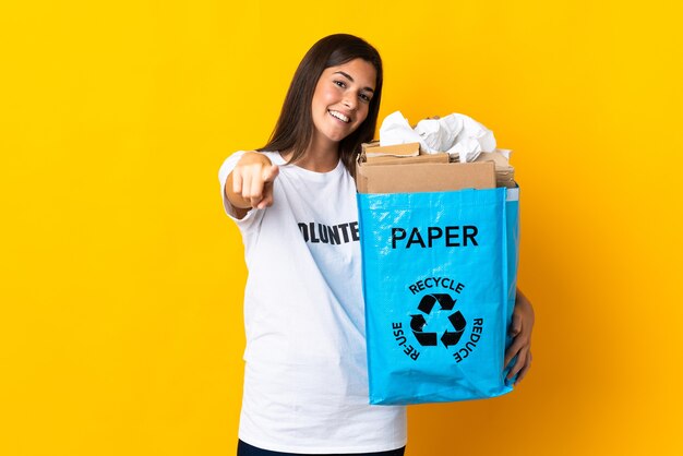 Foto jovem brasileira segurando uma sacola de reciclagem cheia de papel para reciclar na frente amarela apontando com expressão feliz