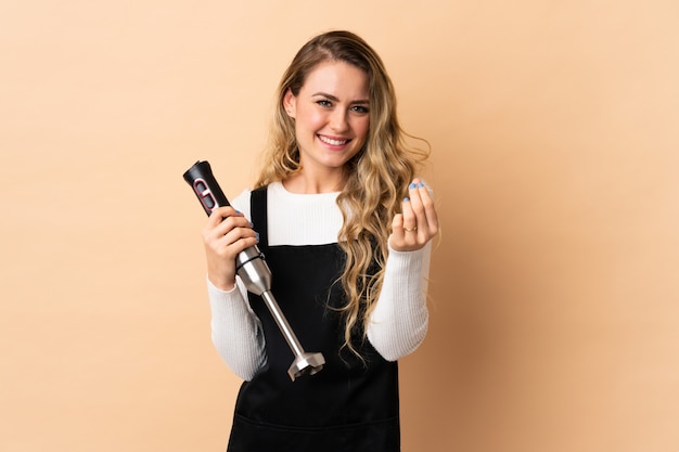 Jovem brasileira usando o liquidificador isolado em bege fazendo dinheiro gesto