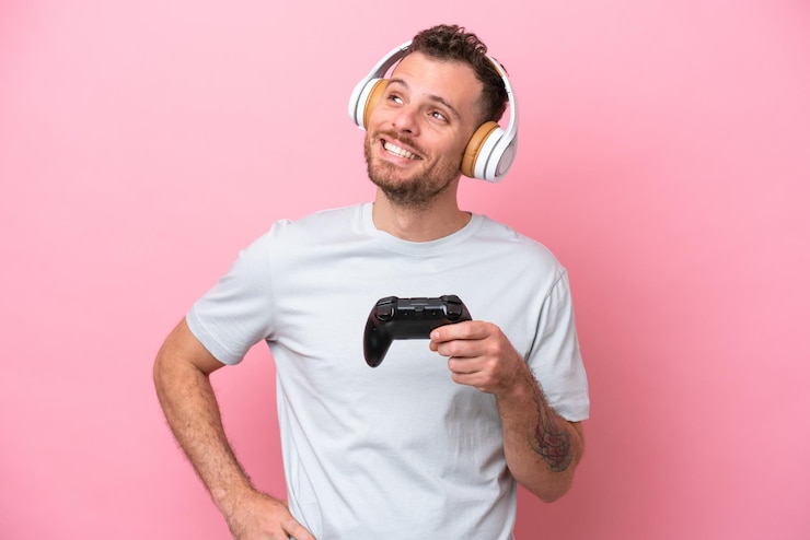 Foto jovem brasileiro brincando com controlador de videogame isolado em fundo rosa posando com braços no quadril e sorrindo