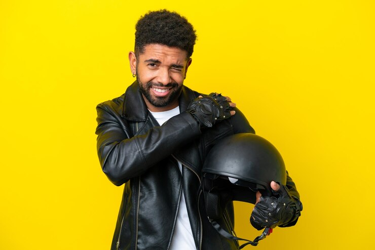 Foto jovem brasileiro com capacete de moto isolado em fundo amarelo sofrendo de dor no ombro por ter feito um esforço