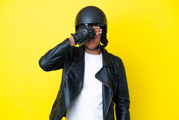 Jovem brasileiro com um capacete de motociclista isolado em um fundo amarelo, cobrindo os olhos com as mãos. Não quero ver nada