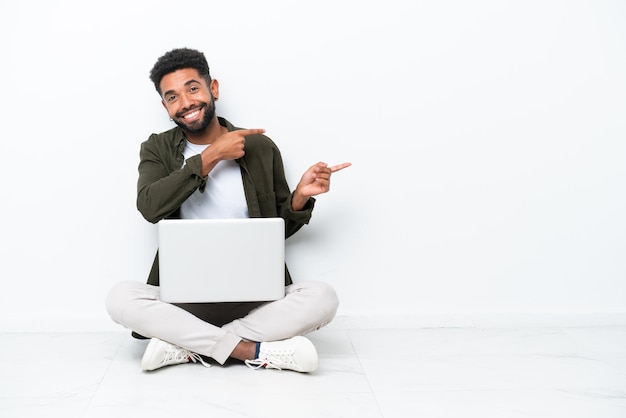 Jovem brasileiro com um laptop sentado no chão isolado em branco apontando o dedo para o lado e apresentando um produto