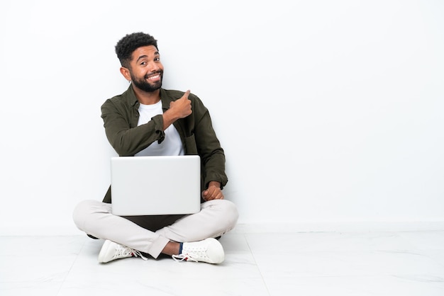 Jovem brasileiro com um laptop sentado no chão isolado em branco apontando para trás