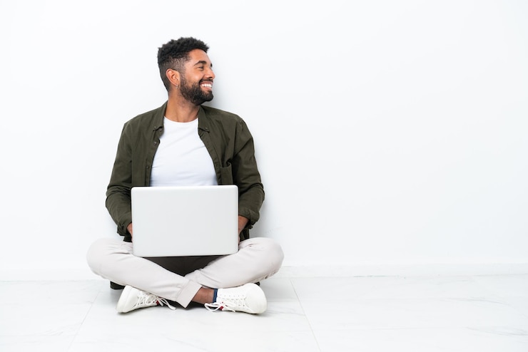 Foto jovem brasileiro com um laptop sentado no chão isolado no branco rindo em posição lateral