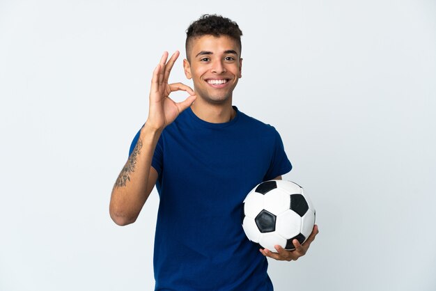 Jovem brasileiro em muro isolado com bola de futebol e fazendo sinal de OK