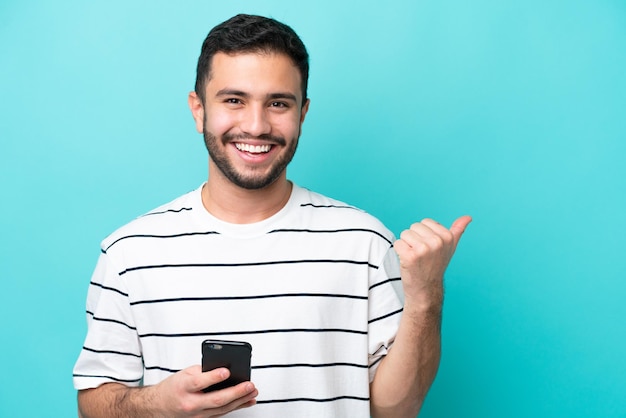 Jovem brasileiro isolado em fundo azul usando telefone celular e apontando para a lateral