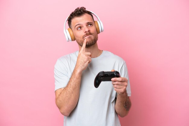 Jovem brasileiro jogando com controlador de videogame isolado em fundo rosa com dúvidas ao olhar para cima