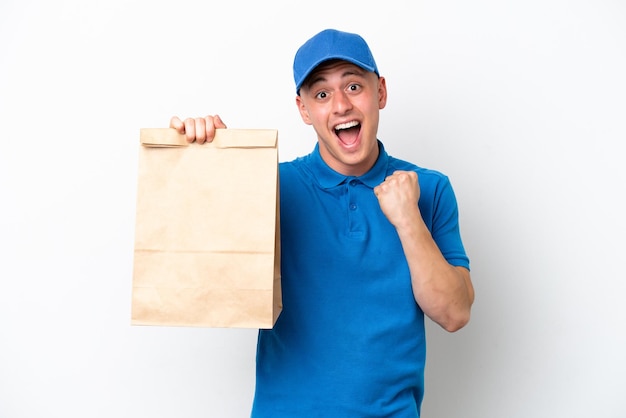 Jovem brasileiro levando um saco de comida para viagem isolado no fundo branco comemorando uma vitória na posição de vencedor