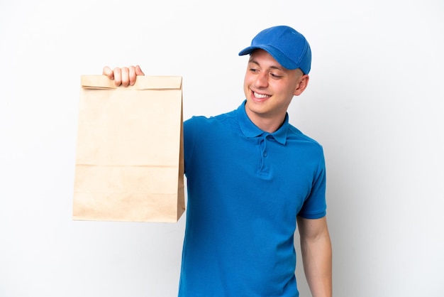 Jovem brasileiro pegando um saco de comida para viagem isolado no fundo branco olhando para o lado e sorrindo