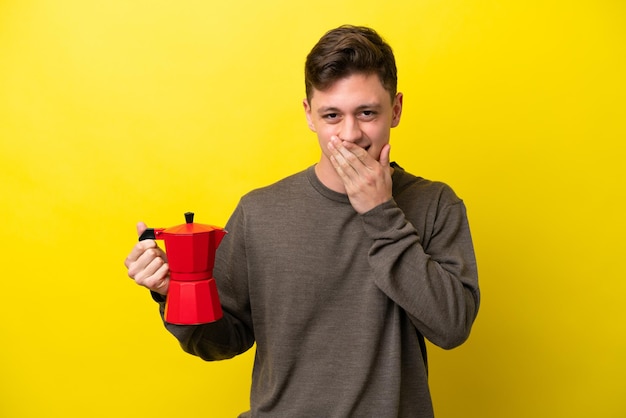 Jovem brasileiro segurando a cafeteira isolada em fundo amarelo feliz e sorridente cobrindo a boca com a mão
