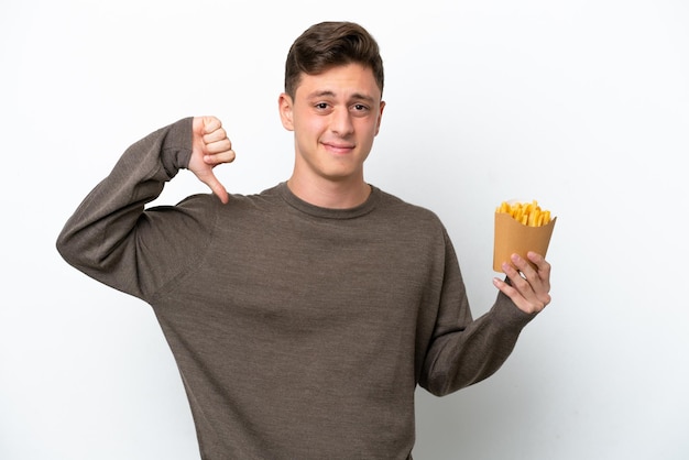 Jovem brasileiro segurando batatas fritas isoladas no fundo branco, mostrando o polegar para baixo com expressão negativa