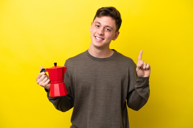 Jovem brasileiro segurando cafeteira isolada em fundo amarelo mostrando e levantando um dedo em sinal do melhor