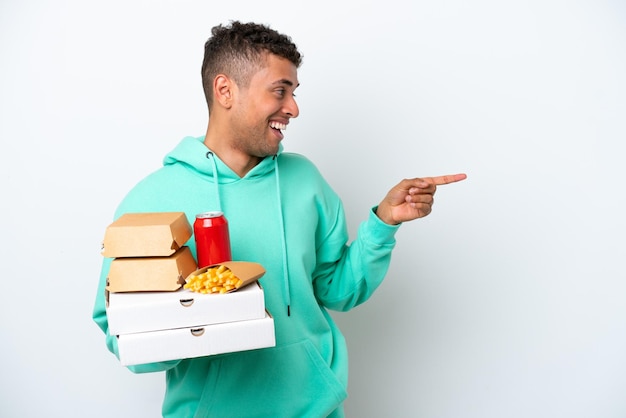Jovem brasileiro segurando fast food isolado no fundo branco, apontando o dedo para o lado e apresentando um produto