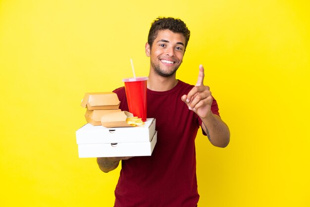 Jovem brasileiro segurando fundo isolado de pizzas e hambúrgueres, mostrando e levantando um dedo