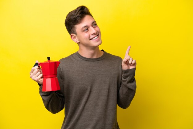 Jovem brasileiro segurando o bule de café isolado em fundo amarelo apontando uma ótima ideia