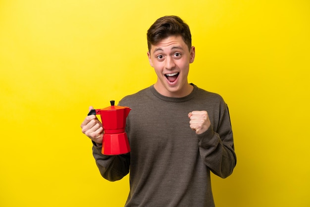 Jovem brasileiro segurando o bule de café isolado em fundo amarelo comemorando uma vitória na posição de vencedor