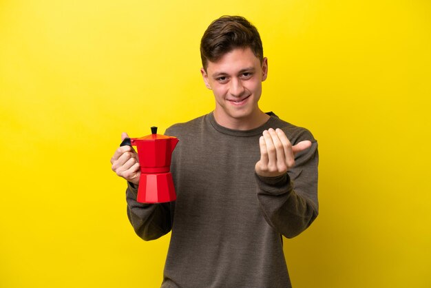 Jovem brasileiro segurando o bule de café isolado em fundo amarelo convidando para vir com a mão feliz por você ter vindo