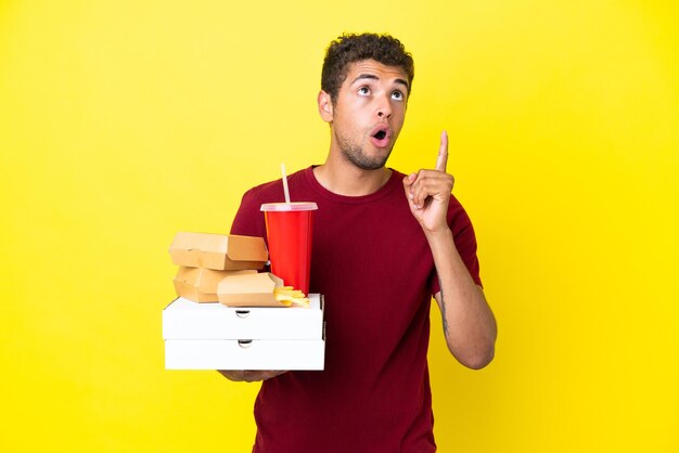 Foto jovem brasileiro segurando pizzas e hambúrgueres isolado de fundo pensando uma ideia apontando o dedo para cima