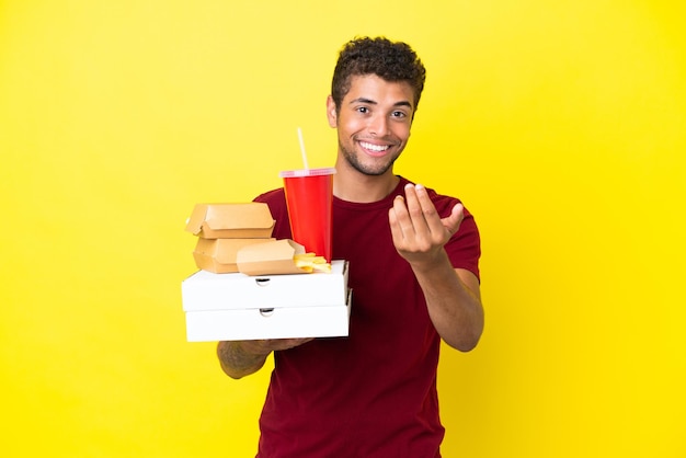 Jovem brasileiro segurando pizzas e hambúrgueres isolados de fundo convidando para vir com a mão Feliz por você ter vindo