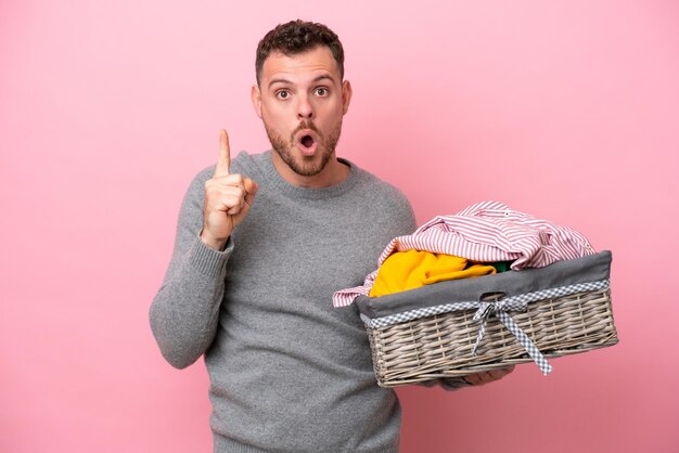 Jovem brasileiro segurando uma cesta de roupas isolada em fundo rosa com a intenção de perceber a solução enquanto levanta um dedo