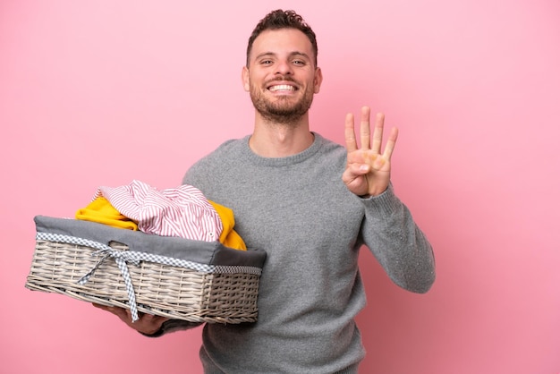 Jovem brasileiro segurando uma cesta de roupas isolada em fundo rosa feliz e contando quatro com os dedos