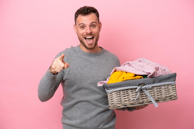 Jovem brasileiro segurando uma cesta de roupas isolada em fundo rosa surpreso e apontando para frente