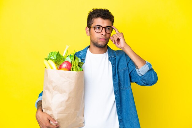 Jovem brasileiro segurando uma sacola de compras de supermercado isolada em um fundo amarelo, tendo dúvidas e pensando