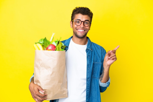 Jovem brasileiro segurando uma sacola de compras isolada em fundo amarelo apontando para o lado para apresentar um produto