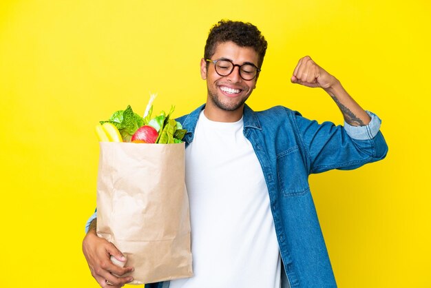 Jovem brasileiro segurando uma sacola de compras isolada em fundo amarelo, fazendo um gesto forte