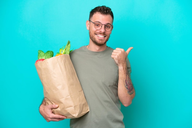 Jovem brasileiro segurando uma sacola de compras isolada em fundo azul apontando para o lado para apresentar um produto