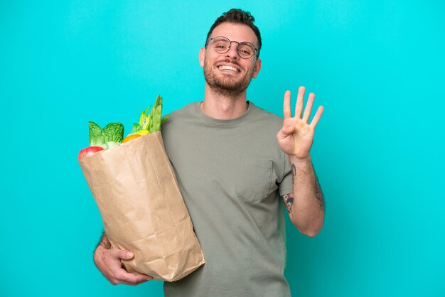 Jovem brasileiro segurando uma sacola de compras isolada em fundo azul feliz e contando quatro com os dedos