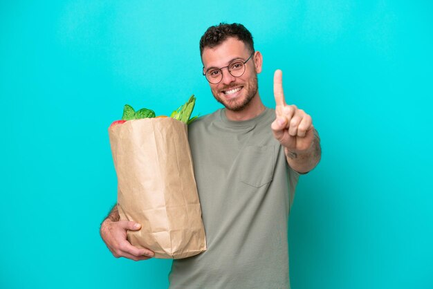 Jovem brasileiro segurando uma sacola de compras isolada em fundo azul mostrando e levantando um dedo