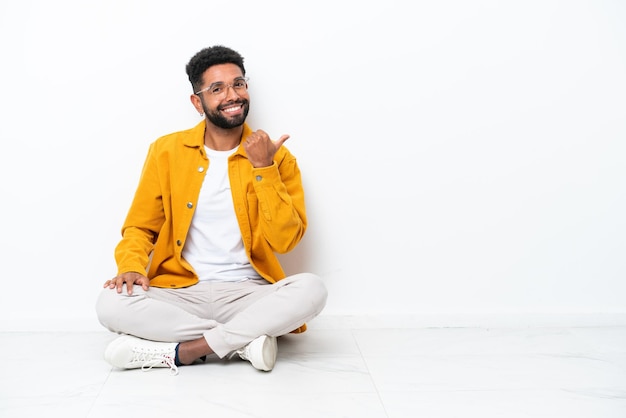 Foto jovem brasileiro sentado no chão isolado no fundo branco apontando para o lado para apresentar um produto