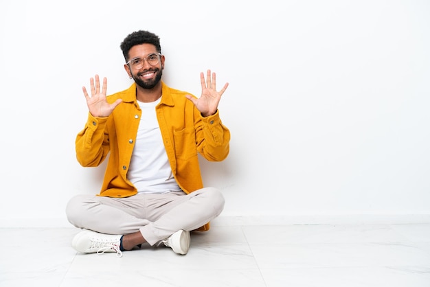 Jovem brasileiro sentado no chão isolado no fundo branco contando dez com os dedos