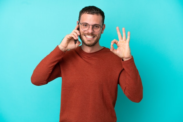 Jovem brasileiro usando celular isolado em fundo azul, mostrando sinal de ok com os dedos