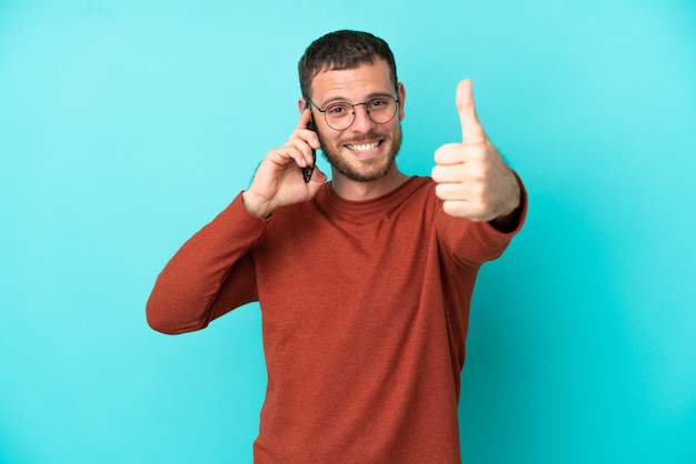 Jovem brasileiro usando telefone celular isolado em um fundo azul com polegar para cima porque algo bom aconteceu