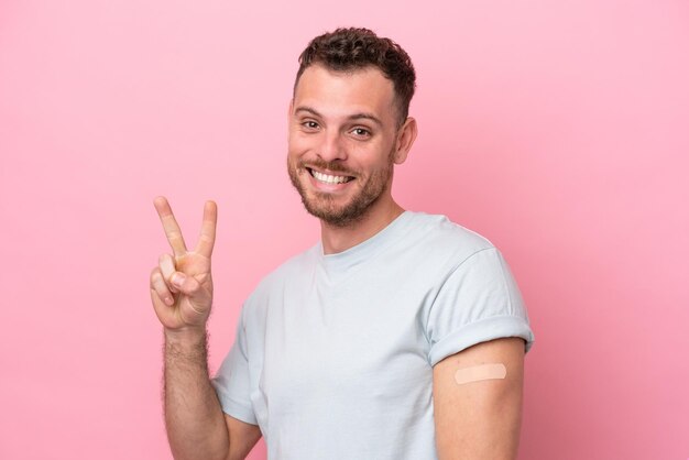Jovem brasileiro vestindo um curativo isolado no fundo rosa sorrindo e mostrando sinal de vitória