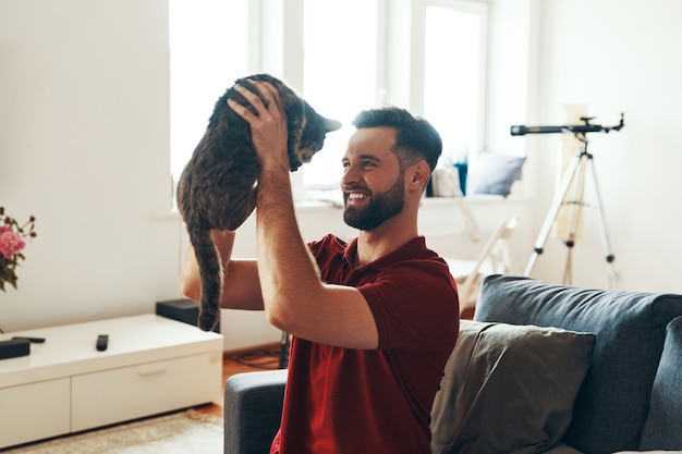 Jovem brincalhão em roupas casuais, criando um vínculo com um gato doméstico e sorrindo enquanto passa um tempo dentro de casa