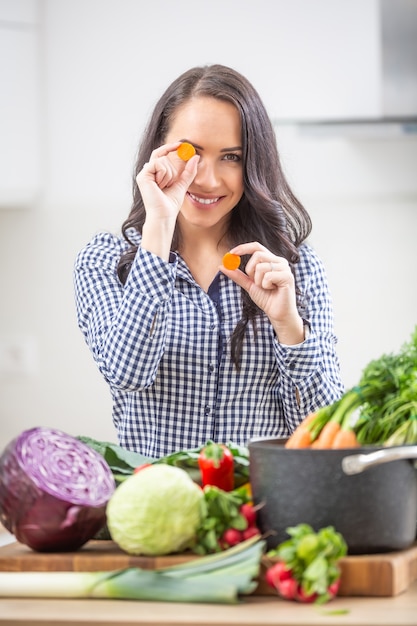 Jovem brincalhão segurando fatias de cenoura na cozinha - dieta vegetal e conceito de saúde.