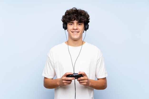 Jovem brincando com um controlador de videogame sobre parede azul isolada, sorrindo muito